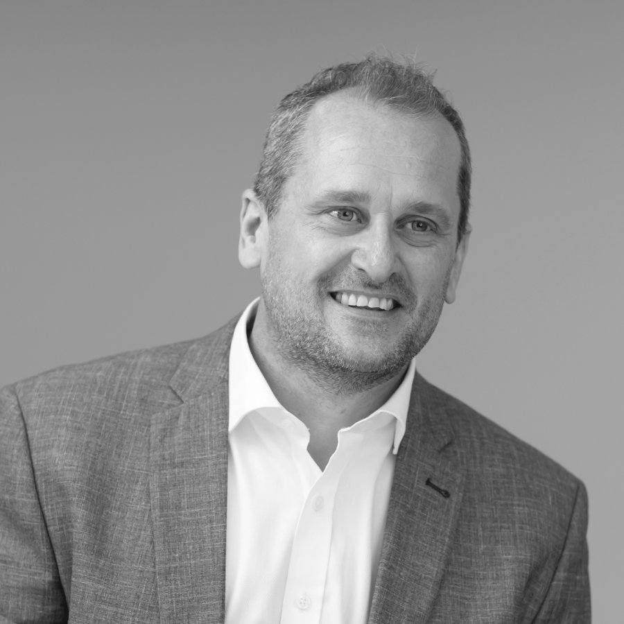 BUSY at Work, or one of the variants): A man with short hair and a neatly trimmed beard, dressed in a suit jacket over a white shirt, smiles as he gazes slightly to the side. The background is plain and neutral. The photograph, facilitated by BUSY at Work, is presented in black and white.