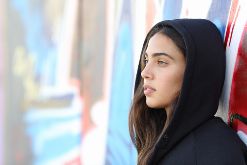Young female in hoodie leans against graffiti wall.