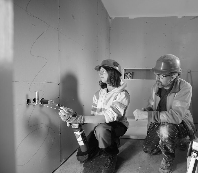 Two construction workers are kneeling in a partially finished room, equipped with hard hats and discussing plans. One of them holds a caulking gun and applies adhesive to a wall panel. The black-and-white photograph captures the essence of women in construction, highlighting their skilled presence in traditionally male-dominated trades, with support by BUSY at Work.