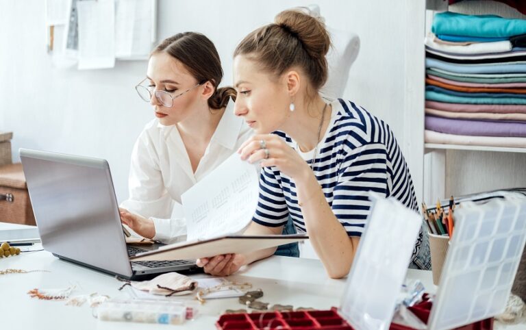 Two women at a desk review a laptop and papers amid sewing materials, exploring job services with support by BUSY at Work.