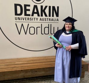 Charley, proudly dressed in a cap and gown, holds a green and blue certificate case in front of the Deakin University Australia Worldly logo. A wooden bench stands nearby, bearing witness to this significant occasion with support by BUSY at Work.