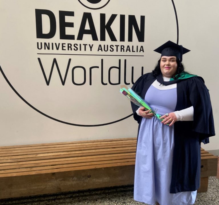 Charley, proudly dressed in a cap and gown, holds a green and blue certificate case in front of the Deakin University Australia Worldly logo. A wooden bench stands nearby, bearing witness to this significant occasion with support by BUSY at Work.