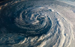 A birds-eye view captures a massive, swirling storm system over the ocean. The storm takes on a spiral shape with thick, white clouds, creating a powerful and dramatic visual against the dark blue waters below.