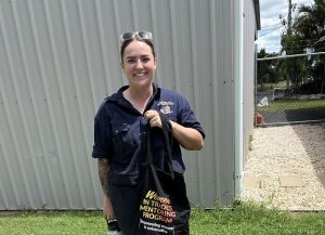 A woman in a blue work shirt is standing outside near a metal building, smiling while holding a black bag that reads Winner in Trades Mentoring Programme, supported by BUSY at Work. They have sunglasses resting on their head, with grass and a chain-link fence visible in the background.