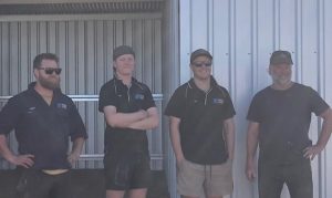 Four men are standing in front of a metal building. Theyre all dressed casually, complete with caps and sunglasses. The background features corrugated metal walls. They seem at ease as they pose for the photograph, with support by BUSY at Work.