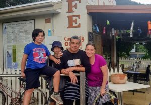 Four people are smiling as they pose in front of a café, where a sign and menu are displayed on the wall. A man stands in the centre, flanked by two boys, while a woman stands on the right holding a bag. Nearby, a bike is parked next to a white picket fence. This was made possible with support from BUSY at Work.