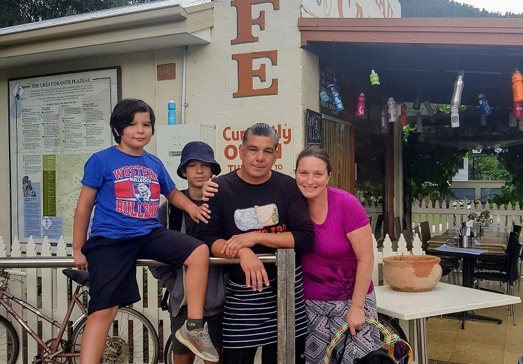 Four people are smiling as they pose in front of a café, where a sign and menu are displayed on the wall. A man stands in the centre, flanked by two boys, while a woman stands on the right holding a bag. Nearby, a bike is parked next to a white picket fence. This was made possible with support from BUSY at Work.