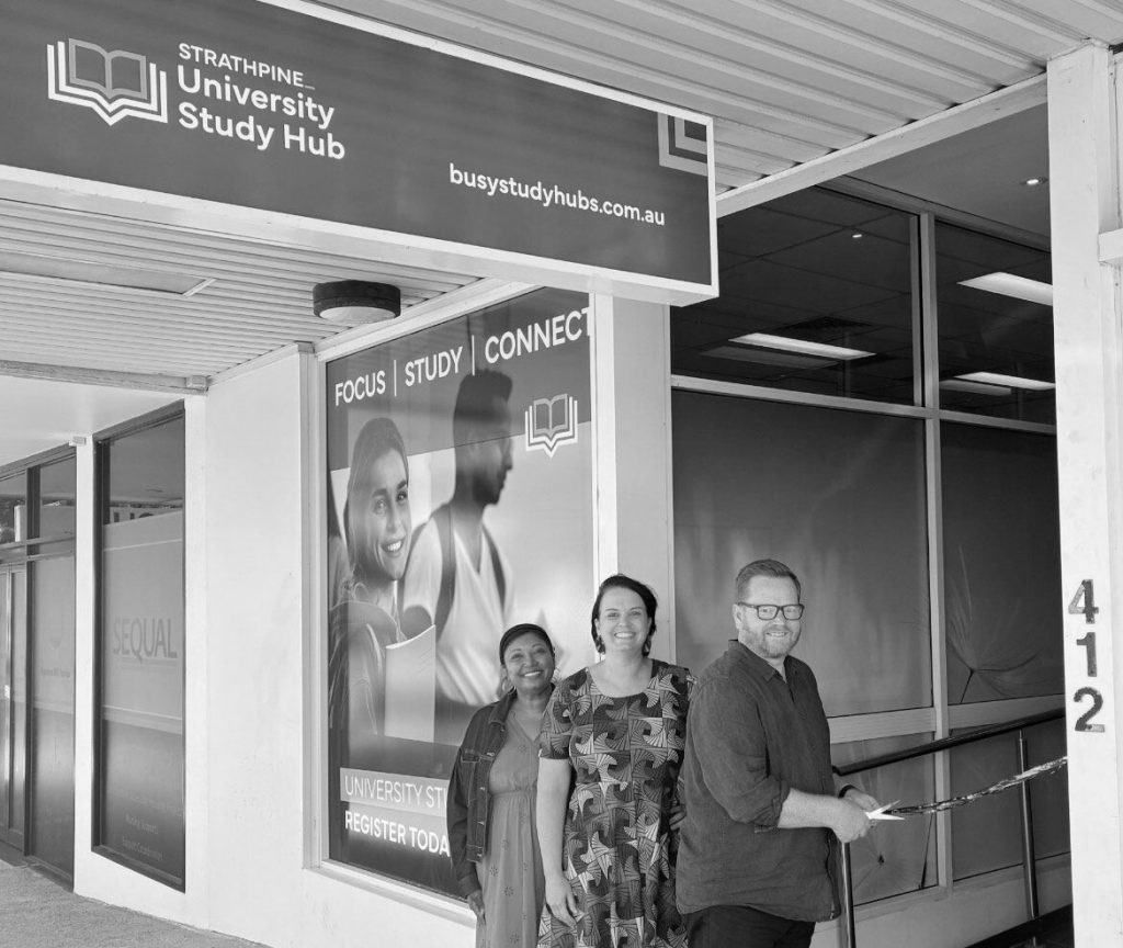 Supported by BUSY at Work, three people are seen smiling in front of the Strathpine University Study Hub. One man is cutting a ribbon with scissors, while two women stand alongside him. Signage on the building features messages related to study.