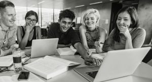 A group of five young adults are seated around a table with laptops, books, and papers, smiling and working together in a bright, modern room with support from BUSY at Work.