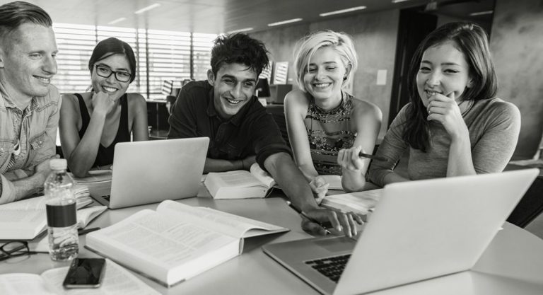 A group of five young adults are seated around a table with laptops, books, and papers, smiling and working together in a bright, modern room with support from BUSY at Work.