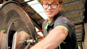 A young man in glasses and a black shirt with green accents operates industrial machinery, with support by BUSY at Work, focused indoors.