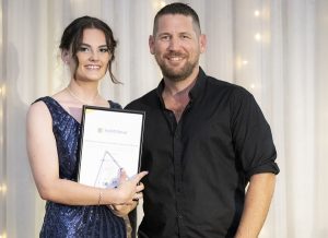 A young woman in a blue dress holds an award with support by BUSY at Work, standing beside a smiling man, in front of lit curtains.