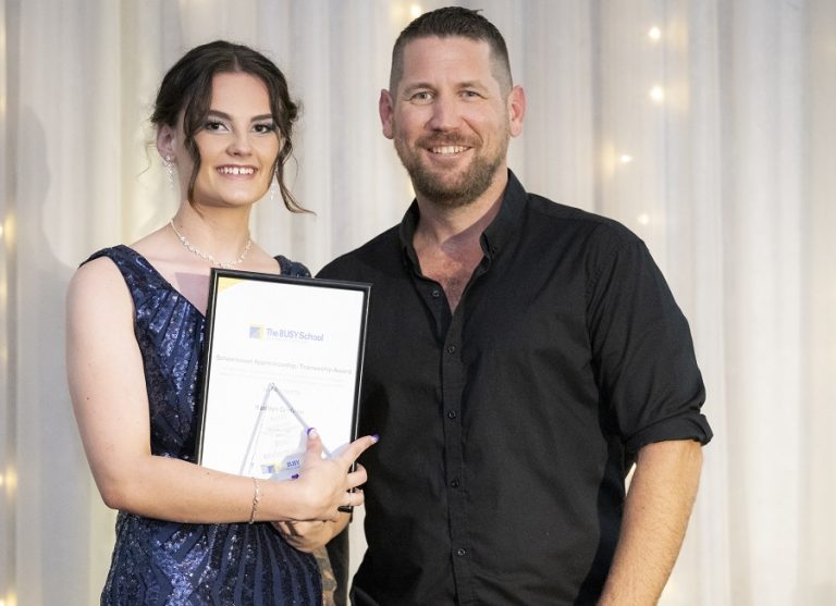 A young woman in a blue dress holds an award with support by BUSY at Work, standing beside a smiling man, in front of lit curtains.