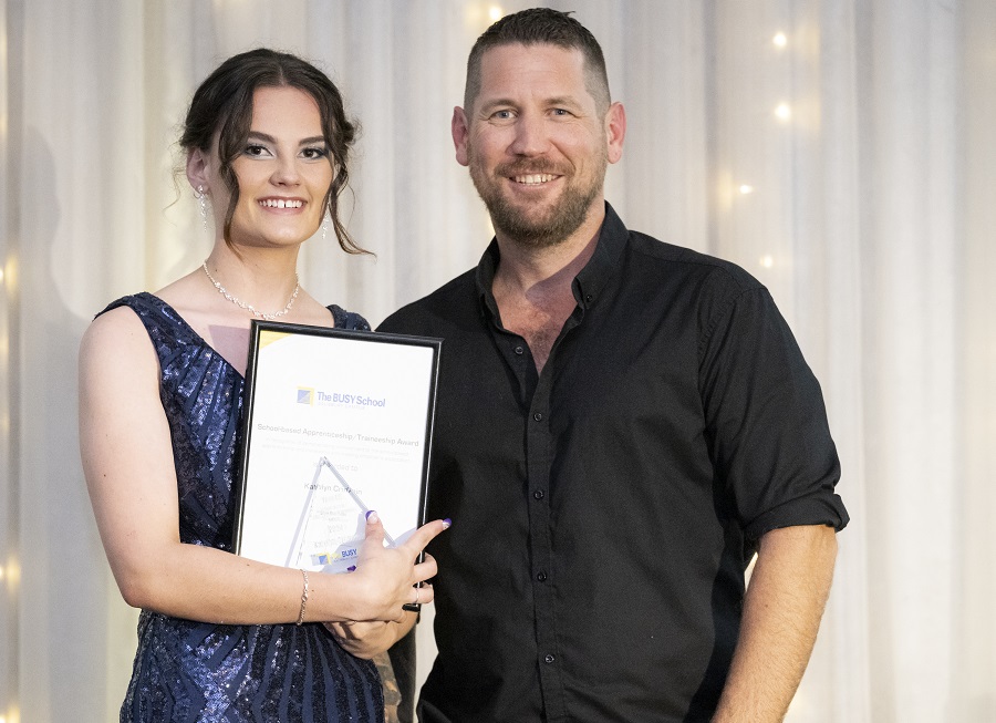A young woman in a blue dress holds an award with support by BUSY at Work, standing beside a smiling man, in front of lit curtains.