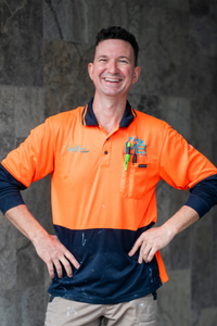 A man in a bright orange and navy work shirt smiles with his hands on his hips before a stone wall, with support by BUSY at Work.