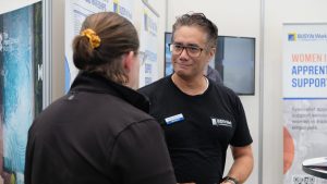 A man in glasses and a BUSY At Work shirt chats with a woman at an expo booth promoting women in apprenticeships, with support by BUSY at Work.