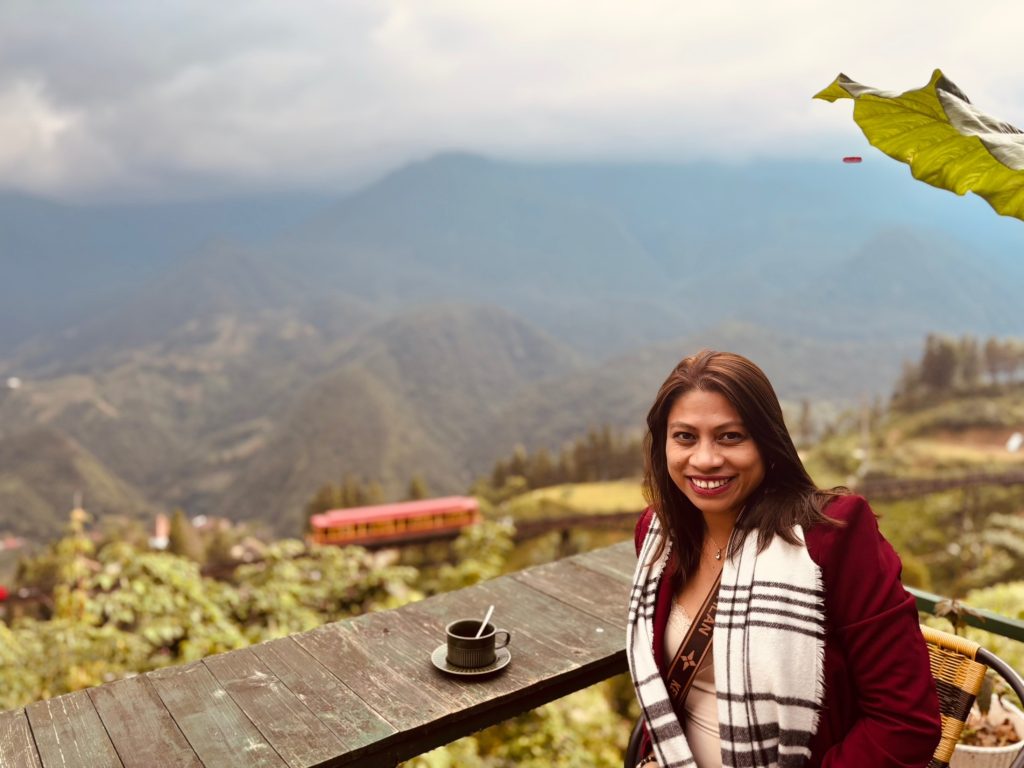 A woman smiling at an outdoor café table with a cup of coffee, enjoying lush mountains and cloudy skies, with support by BUSY at Work.