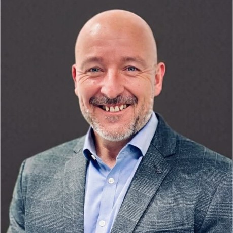 A smiling bald man with a short beard in a grey checked suit jacket and light blue shirt, with support by BUSY at Work, posing against a dark background.