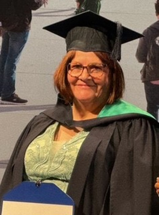 A woman with glasses, a black graduation cap, and a green-sashed gown smiles indoors, with support by BUSY at Work; people blurred behind.