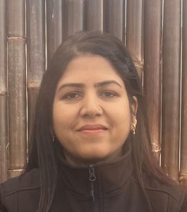 A woman with long dark hair, small gold earrings and a black zip-up jacket stands before bamboo stalks, smiling gently, with support by BUSY at Work.