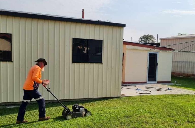 With support by BUSY at Work, a person in an orange long-sleeve shirt mows grass near a cream-coloured building with black-trimmed windows.