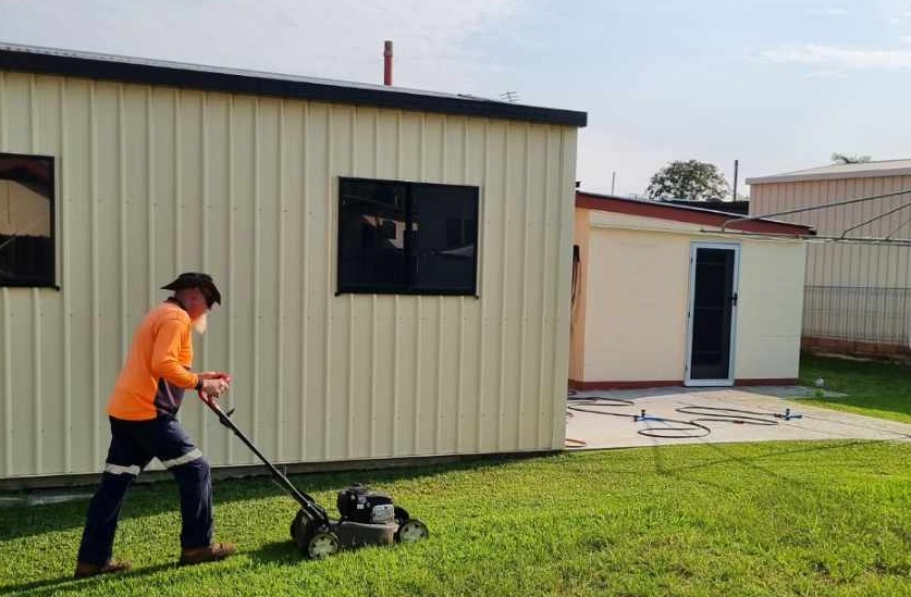 With support by BUSY at Work, a person in an orange long-sleeve shirt mows grass near a cream-coloured building with black-trimmed windows.