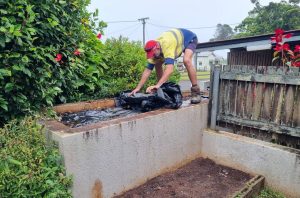 With support by BUSY at Work, a person in a yellow shirt and red cap covers a raised garden bed with black plastic near a wooden fence.