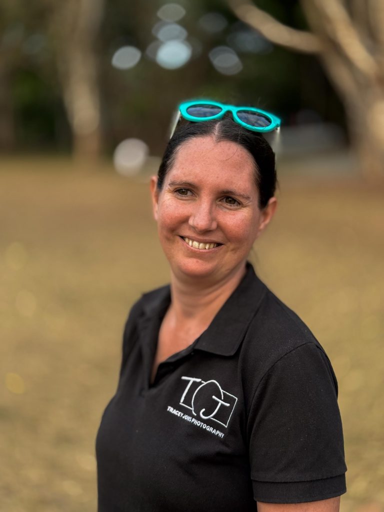 A woman with dark hair tied back, turquoise sunglasses on her head and a TGJ PHOTOGRAPHY shirt, smiling outdoors with support by BUSY at Work.
