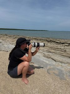 With help and support by BUSY at Work, a person in a black shirt and grey shorts kneels barefoot on a rocky beach, photographing the ocean.