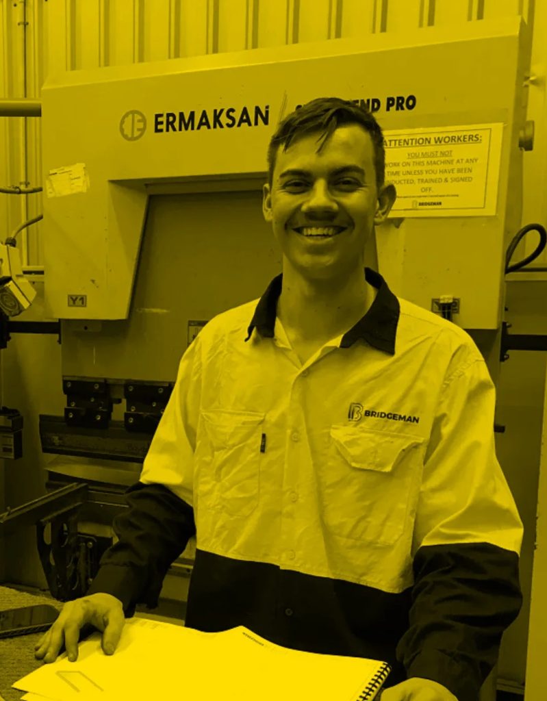 A smiling person in a Bridgeman uniform, with support by BUSY at Work, holds documents by an ERMAKSAN machine in a yellow-tinted workshop.