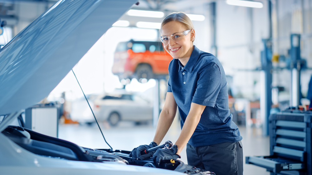 A smiling mechanic wearing safety glasses and a navy polo works under a car bonnet in a bright workshop, with support by BUSY at Work.