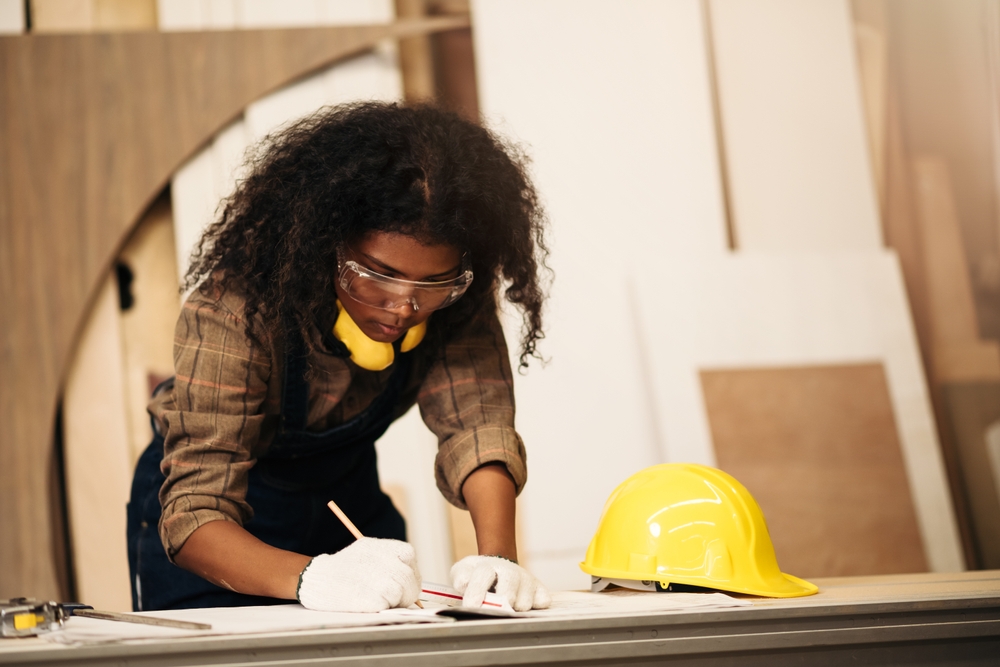 A person in safety glasses, gloves and a plaid shirt draws at a table, with support by BUSY at Work. Hard hat and ear muffs nearby.