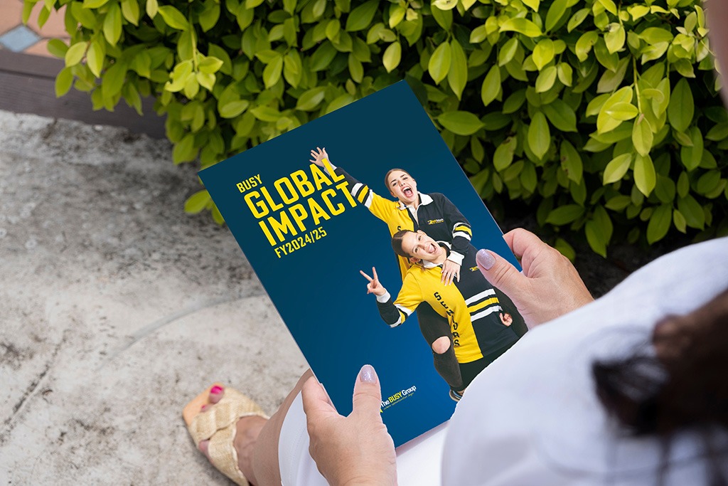 Outdoors, a person holds a Global Impact FY2024/25 brochure, with support by BUSY at Work. Green leaves and two smiling people appear.