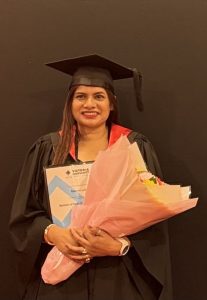 A woman in a graduation cap and gown smiles, holding flowers and a certificate, with support by BUSY at Work, against a dark backdrop.