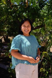 A woman with long dark hair, in a green and white checked blouse and light trousers, smiles outdoors in a sunny garden with support by BUSY at Work.