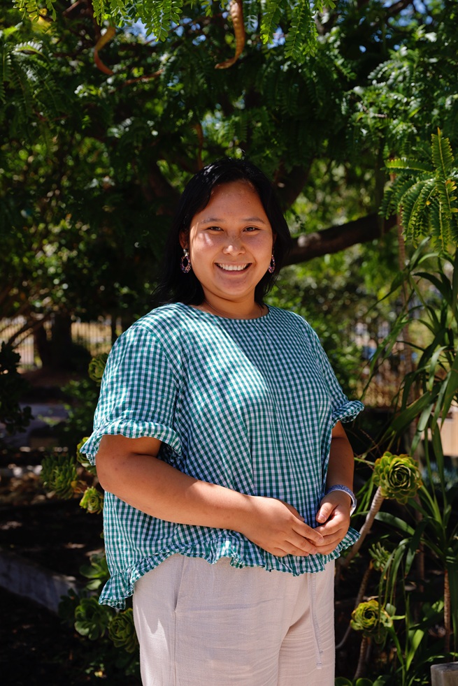 A woman with long dark hair, in a green and white checked blouse and light trousers, smiles outdoors in a sunny garden with support by BUSY at Work.