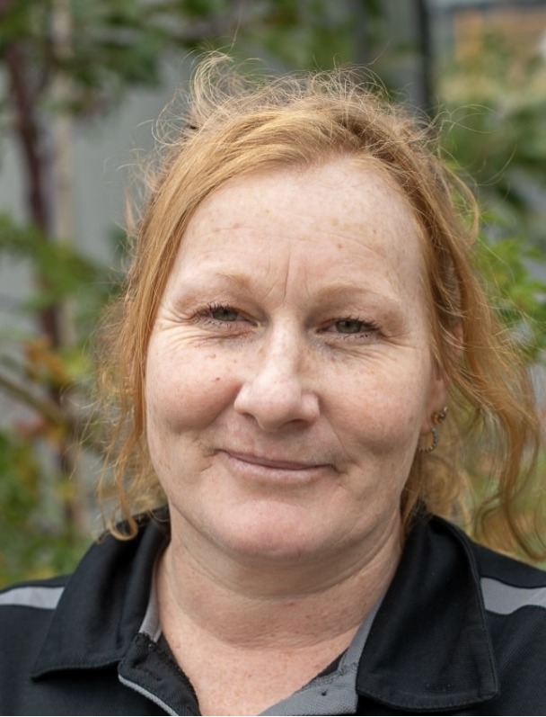 A middle-aged woman with fair skin and wavy reddish-blonde hair smiles gently, with support by BUSY at Work. Greenery is blurred behind.