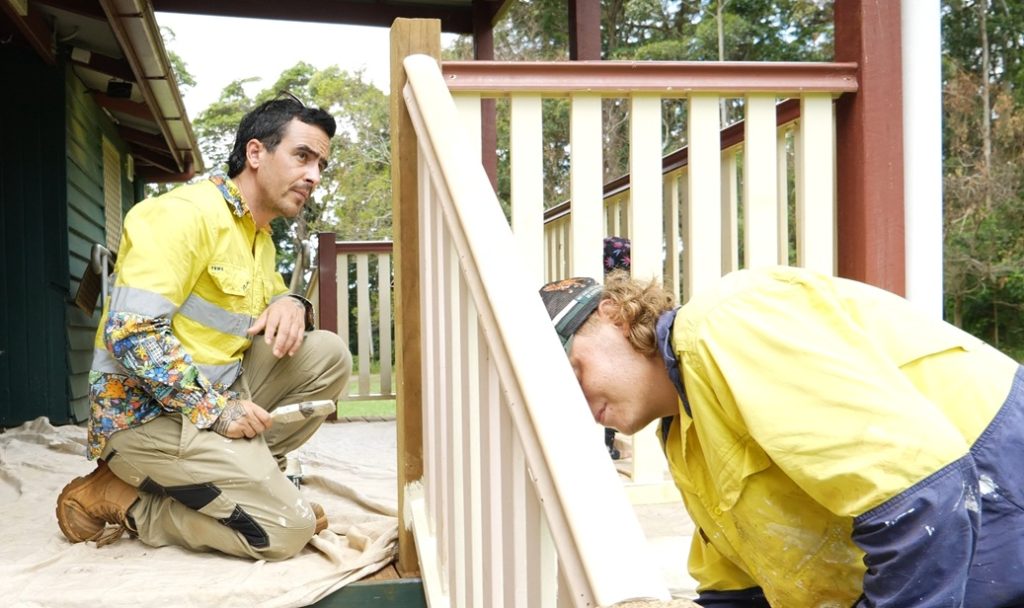 With support from BUSY at Work, two people in yellow work shirts paint a wooden railing outside, with trees and a house in the background.