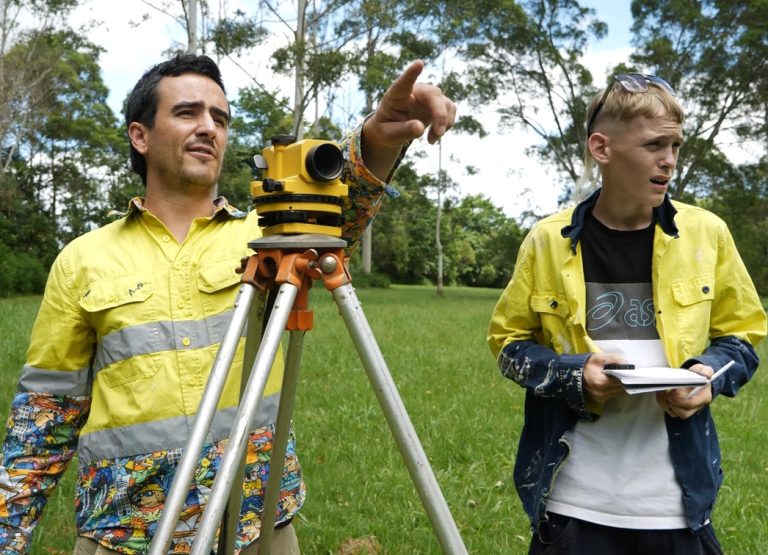 With support by BUSY at Work, two people in yellow work shirts use a surveying tripod in a grassy area with trees behind them.