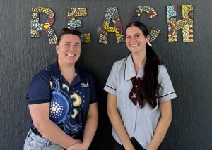 Two young women, smiling in front of a dark wall with “REACH” in colourful mosaic letters, with support by BUSY at Work.