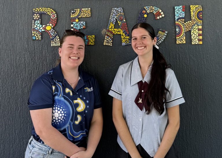 Two young women, smiling in front of a dark wall with “REACH” in colourful mosaic letters, with support by BUSY at Work.