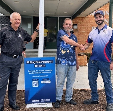 Three men stand outside a building; two shake hands and smile near a blue 'Skilling Queenslanders for Work' sign, with support by BUSY at Work.