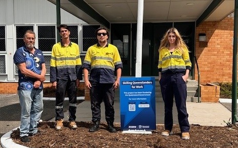 Four people, with support by BUSY at Work, stand outside near a Skilling Queenslanders for Work sign; three wear yellow hi-vis shirts.