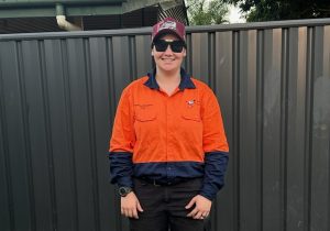 A person in a red and navy hi-vis work shirt, black trousers, maroon cap, and sunglasses smiles with support by BUSY at Work.