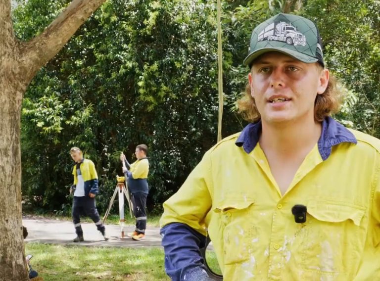 A man in a yellow and blue work shirt and green cap speaks to camera, with support by BUSY at Work at Mt Tamborine Construction Programme.