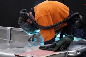A person in protective gear, including a welding helmet and respirator, welds metal with blue sparks, supported by BUSY at Work; gloves and clipboard nearby.