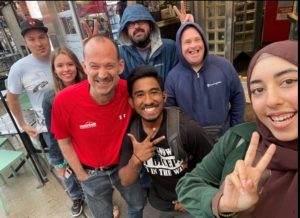 Seven people smile and flash peace signs in a rainy outdoor selfie, with help and support by BUSY at Work. Hoodies and jackets keep them dry.
