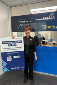 A woman in a black uniform smiles, holding a Skilling Queenslanders for Work sign with support by BUSY at Work outside Workforce Australia.