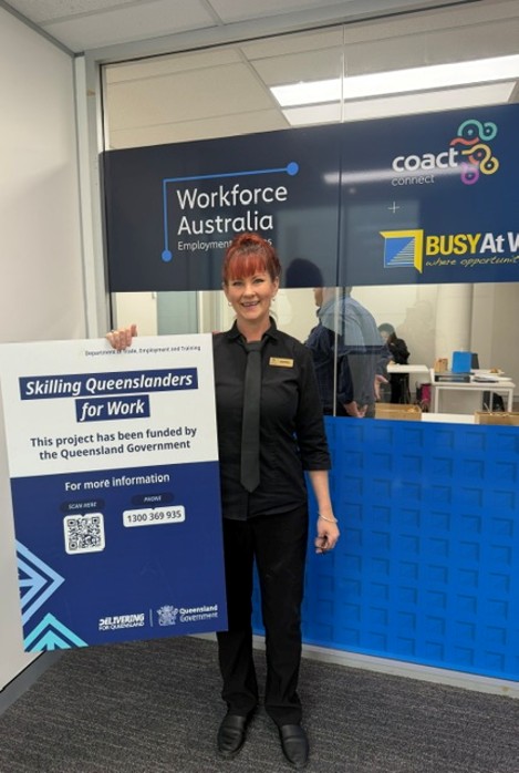 A woman in a black uniform smiles, holding a Skilling Queenslanders for Work sign with support by BUSY at Work outside Workforce Australia.